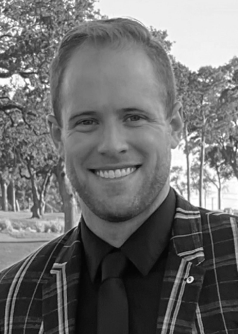 black and white image of a smiling man with blonde hair wearing a dark button up shirt with a dark tie and dark blazer with light  stripes