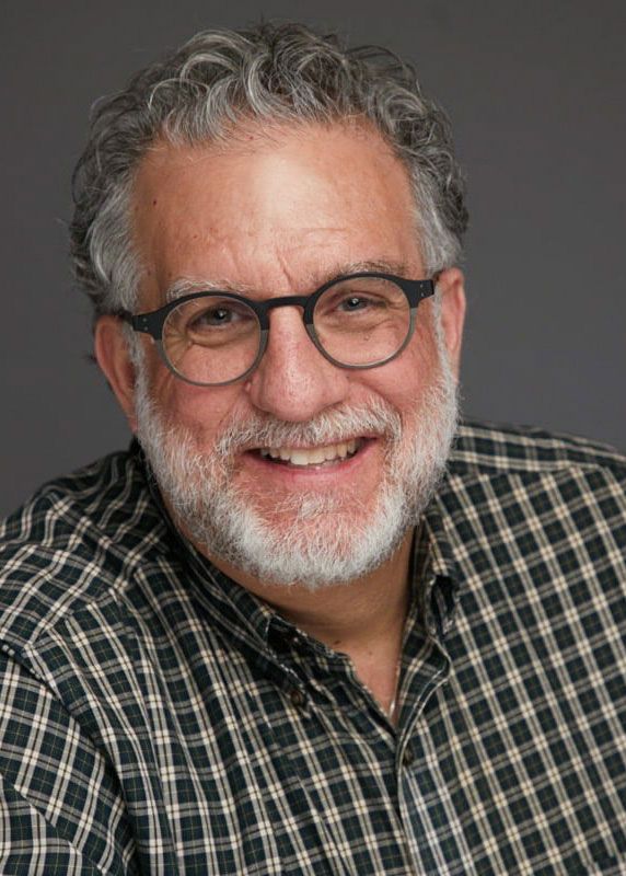 image oa a smiling man with salt and pepper curly hair and a gray beard wearing black round eyeglasses and a checkered black and yellow button up shirt