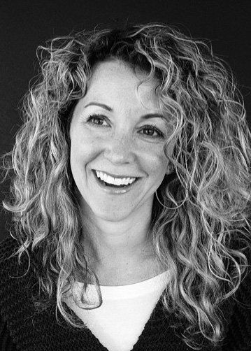 black and white image of a smiling woman with a lot of curly blonde hair wearing a white t-shirt and a black v-neck sweater
